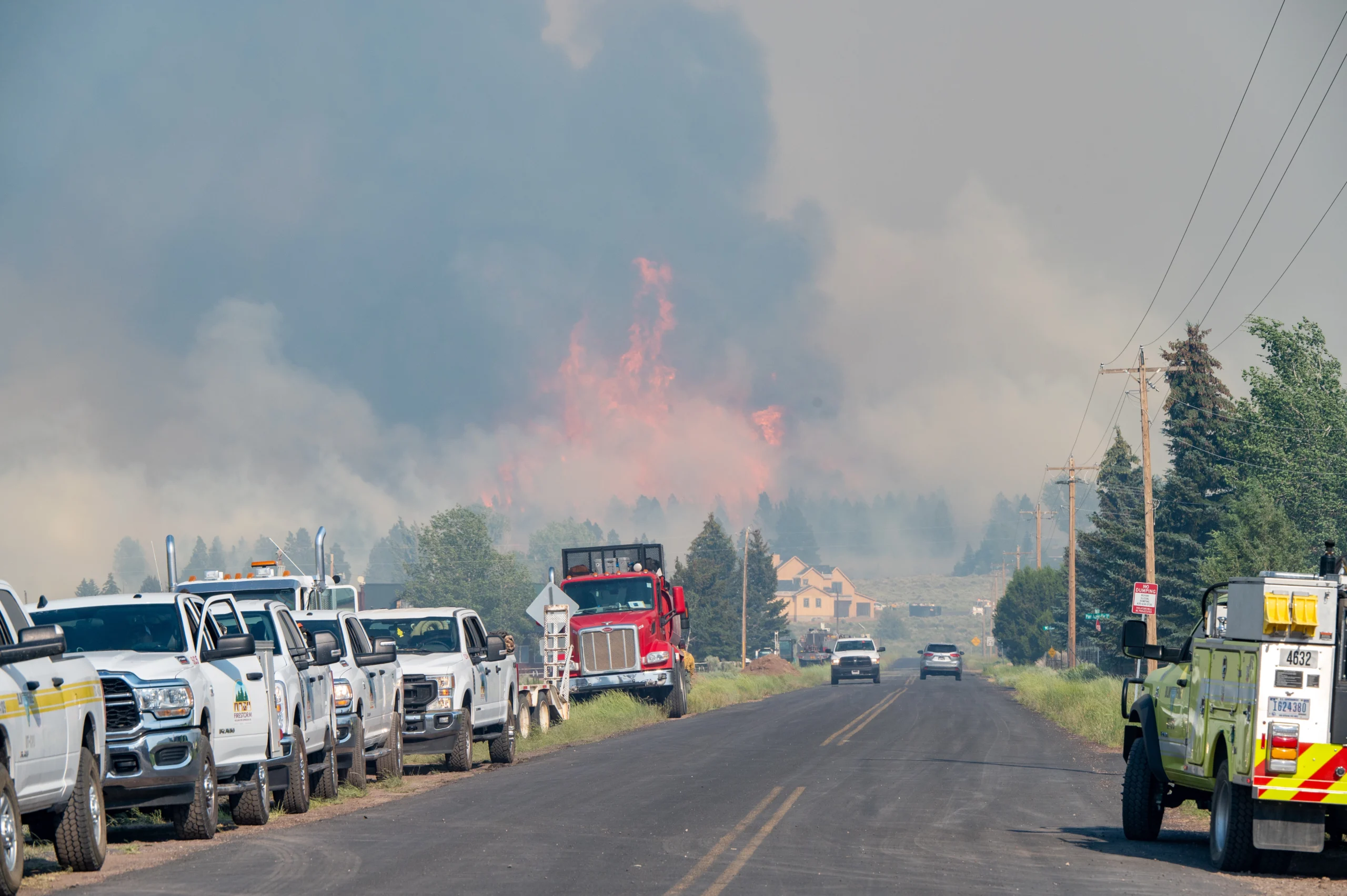 A line of work trucks parked on a road facing a massive wildfire and thick smoke.