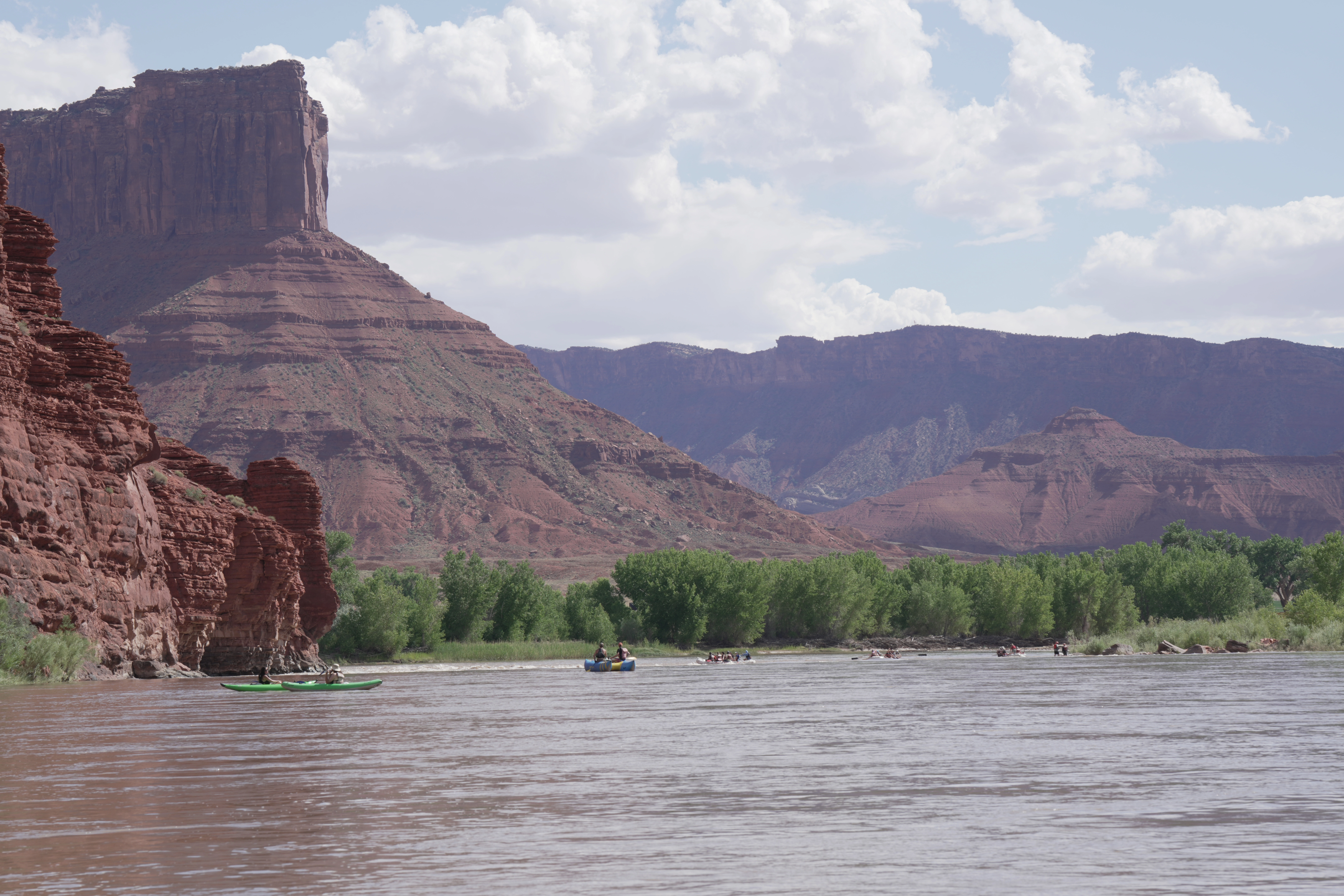 People in kayaks and rafts on a river below towering red rock cliffs