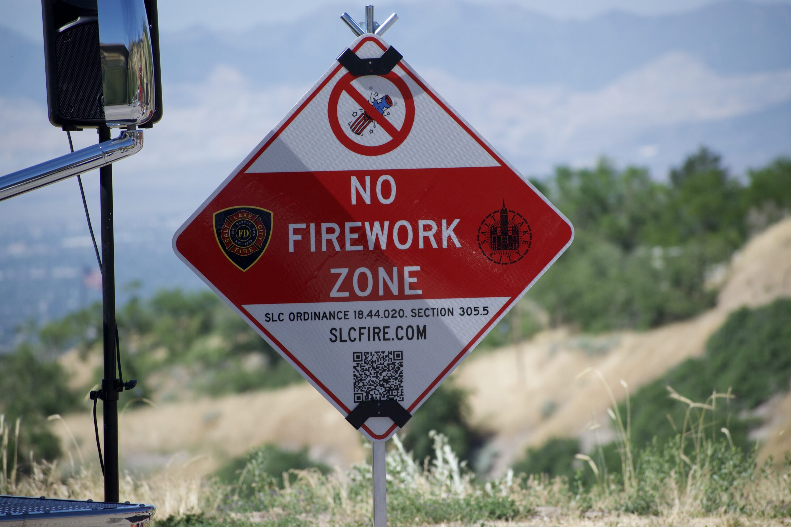 Red “No Firework Zone” sign with a QR code and city ordinance info, outdoors by a hill.
