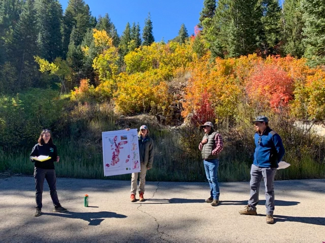 Four people stand on an asphalt path in front of trees with colorful autumn foliage
