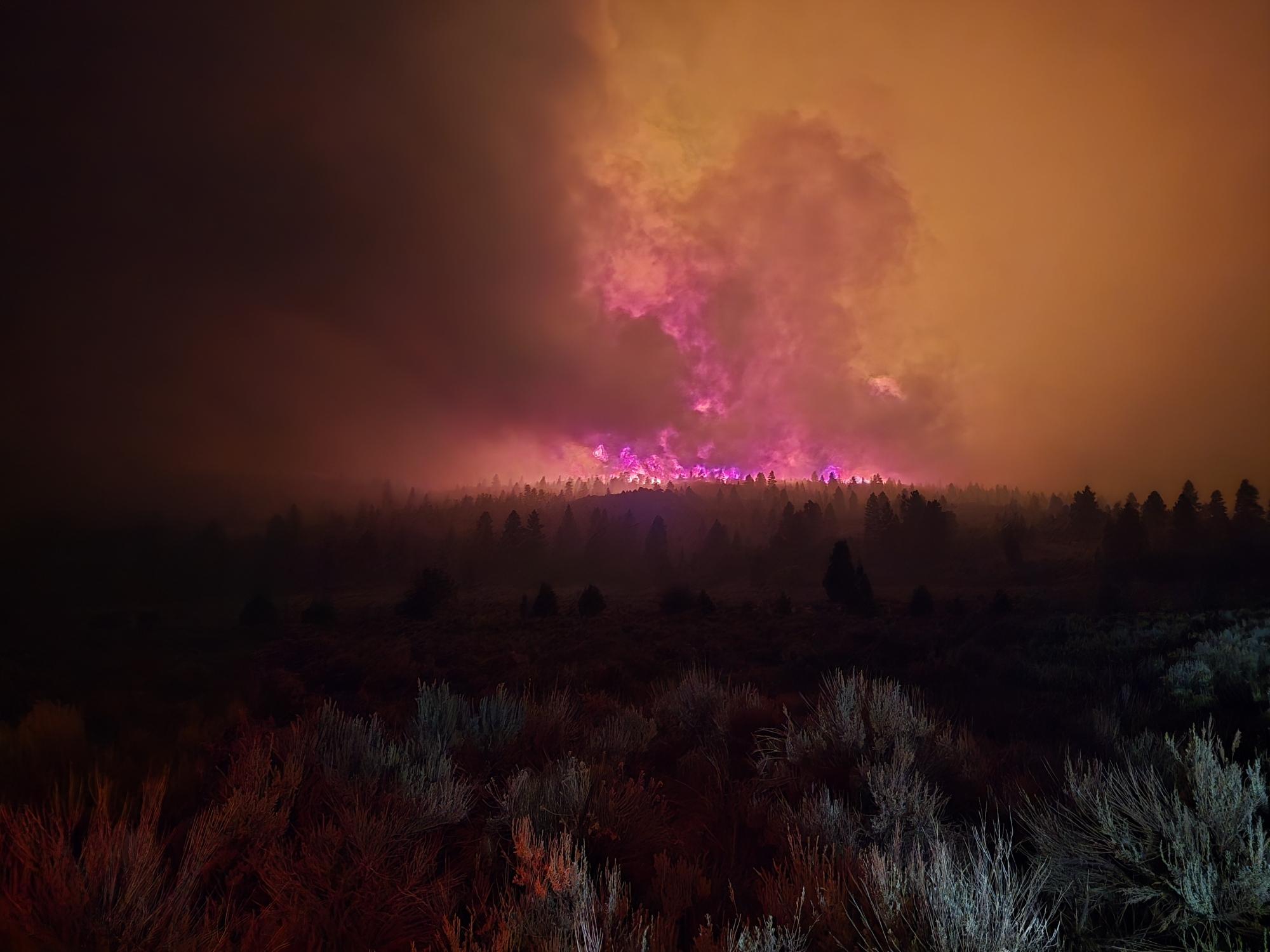 Massive smoke plume glowing red from a wildfire, rising above a dark forest silhouette at night.