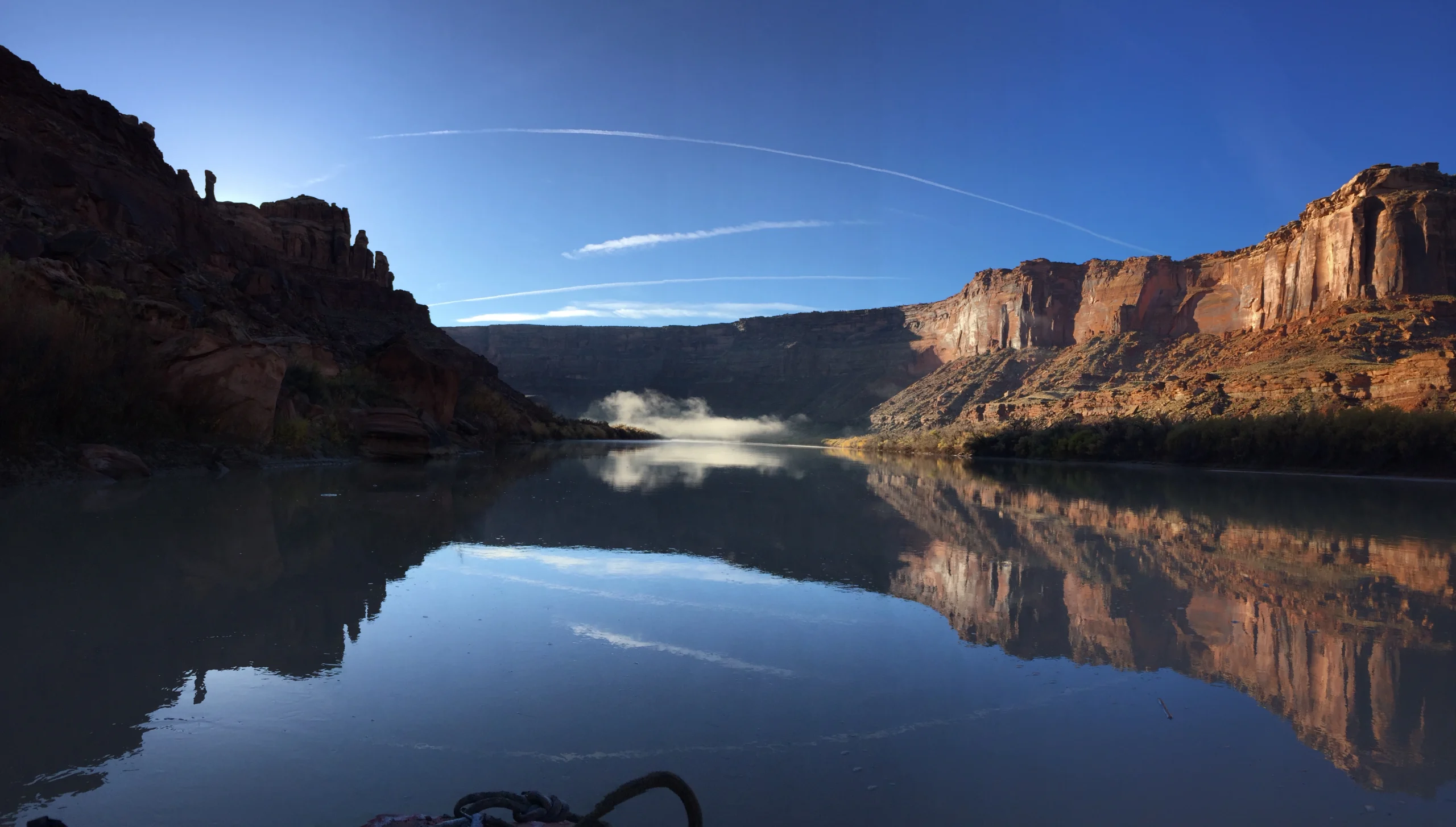 Wide view of a river canyon with red cliffs, perfect reflections, and distant mist
