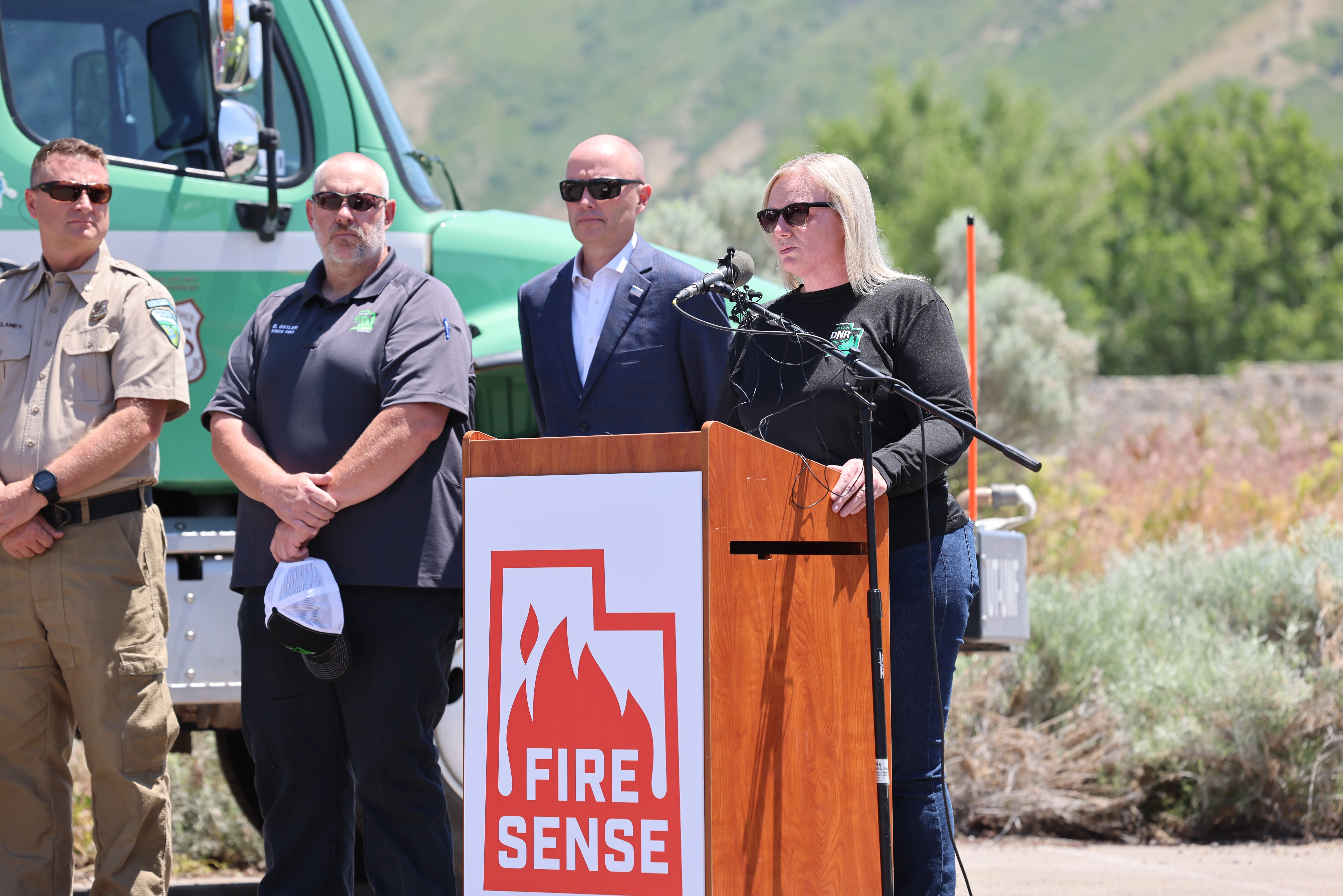 A woman speaks at a "FIRE SENSE" podium with three men standing behind her.